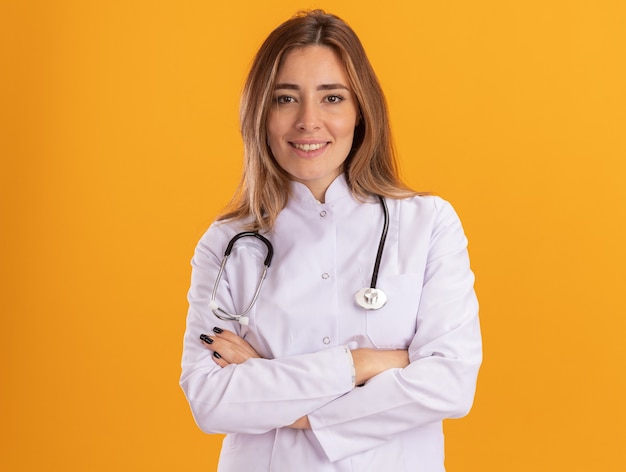 노란색 벽에 고립 된 청진 교차 손으로 의료 가운을 입고 웃는 젊은 여성 의사
