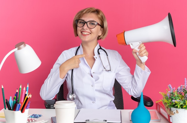 Sorridente giovane dottoressa che indossa accappatoio medico con stetoscopio e occhiali si siede alla scrivania con strumenti medici che tengono e punta all'altoparlante isolato su sfondo rosa