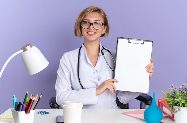 청진기와 안경을 쓴 의료 가운을 입은 웃고 있는 젊은 여성 의사는 파란색 배경에 격리된 클립보드를 들고 의료 도구를 들고 테이블에 앉아 있습니다.