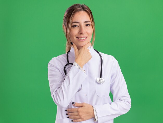 Smiling young female doctor wearing medical robe with stethoscope grabbed chin isolated on green wall