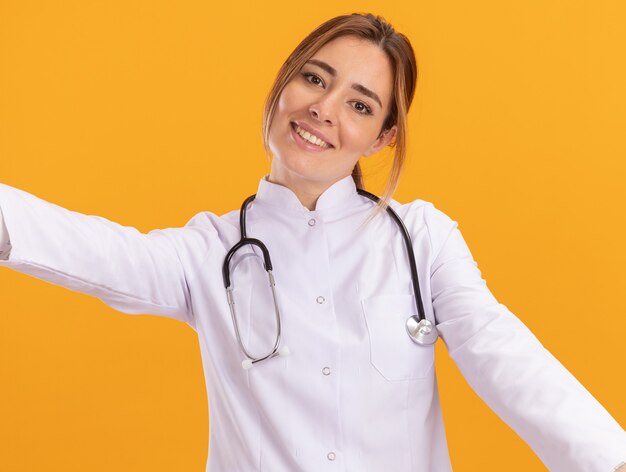 Smiling young female doctor wearing medical robe with stethoscope holding front isolated on yellow wall