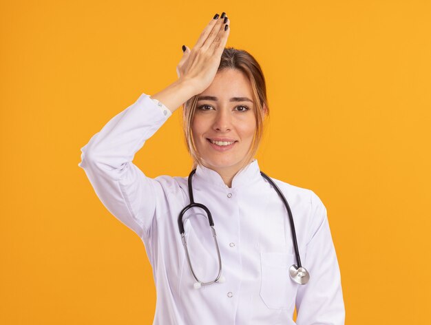 Улыбающаяся молодая женщина-врач в медицинском халате со стетоскопом, держащая руку на храме, изолированном на желтой стене