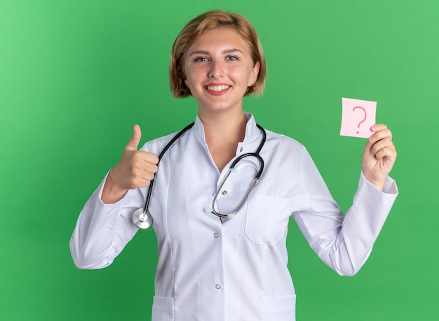 Smiling young female doctor wearing medical robe with stethoscope holding paper question note showing thumb up isolated on green background