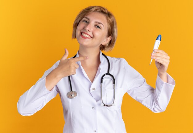 Улыбающаяся молодая женщина-врач в медицинском халате со стетоскопом и указывает на термометр, изолированные на оранжевом фоне