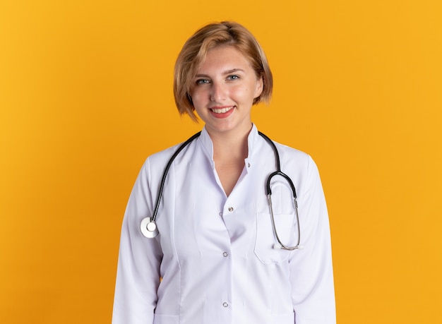 Smiling young female doctor wearing medical robe with stethoscope isolated on orange background