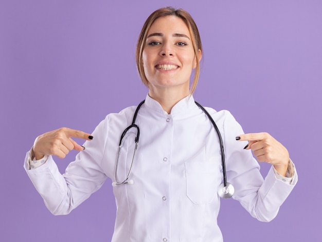 Улыбающаяся молодая женщина-врач в медицинском халате со стетоскопом указывает на камеру, изолированную на фиолетовой стене