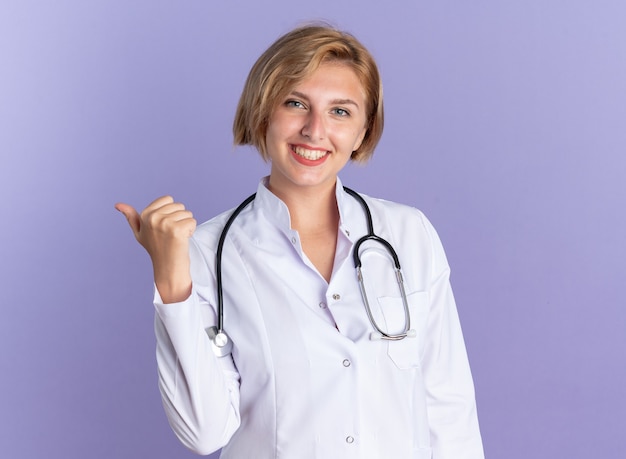 Smiling young female doctor wearing medical robe with stethoscope points at side isolated on blue background with copy space