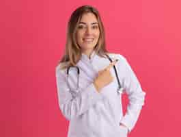 Free photo smiling young female doctor wearing medical robe with stethoscope points at side isolated on pink wall with copy space