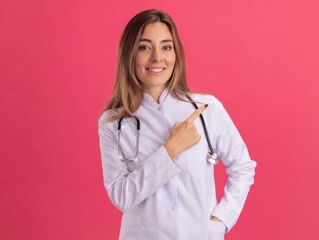 Улыбающаяся молодая женщина-врач в медицинском халате со стетоскопом указывает сбоку, изолированную на розовой стене с копией пространства