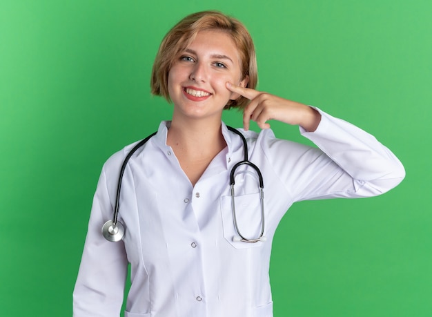 청진기가 녹색 배경에 격리된 뺨에 손가락을 대고 의료 가운을 입은 웃고 있는 젊은 여성 의사