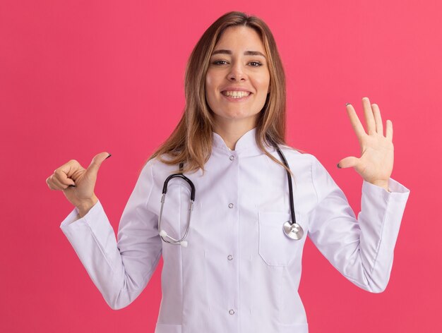ピンクの壁に分離されたさまざまなジェスチャーを示す聴診器で医療ローブを着て笑顔の若い女性医師