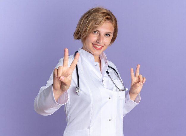 파란색 배경에 고립 된 평화 제스처를 보여주는 청진 기 의료 가운을 입고 웃는 젊은 여성 의사