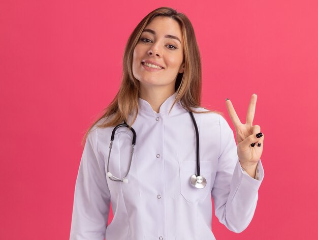 분홍색 벽에 고립 된 평화 제스처를 보여주는 청진 기 의료 가운을 입고 웃는 젊은 여성 의사