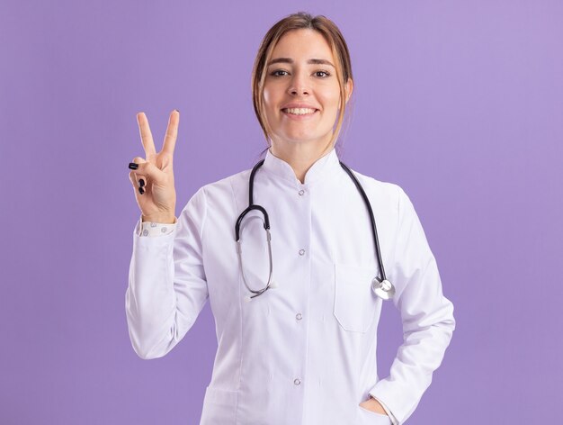 紫色の壁に分離された平和のジェスチャーを示す聴診器で医療ローブを着て笑顔の若い女性医師