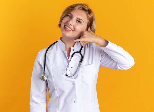 Sorridente giovane dottoressa che indossa abito medico con stetoscopio che mostra gesto di telefonata isolato su sfondo arancione