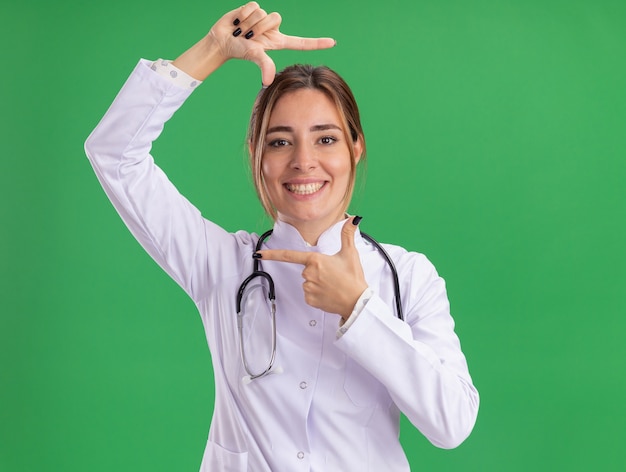 녹색 벽에 고립 된 사진 제스처를 보여주는 청진 기 의료 가운을 입고 웃는 젊은 여성 의사