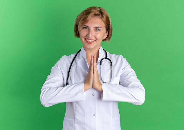 Smiling young female doctor wearing medical robe with stethoscope showing pray gesture isolated on green background