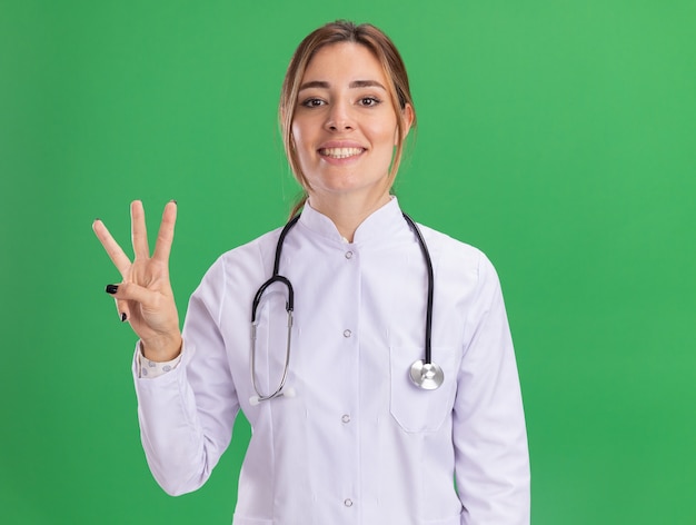 Улыбающаяся молодая женщина-врач в медицинском халате со стетоскопом показывает три изолированные на зеленой стене