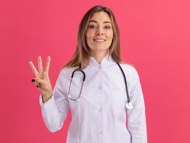 Smiling young female doctor wearing medical robe with stethoscope showing three isolated on pink wall