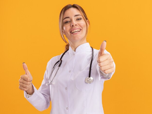 Улыбающаяся молодая женщина-врач в медицинском халате со стетоскопом показывает палец вверх изолированной на желтой стене