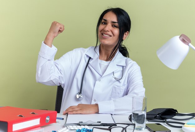 Free photo smiling young female doctor wearing medical robe with stethoscope sits at desk with medical tools showing yes gesture isolated on olive green background