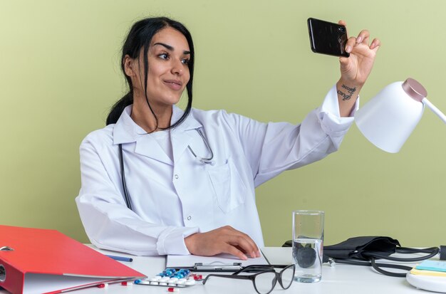 Smiling young female doctor wearing medical robe with stethoscope sits at desk with medical tools take a selfie isolated on olive green background