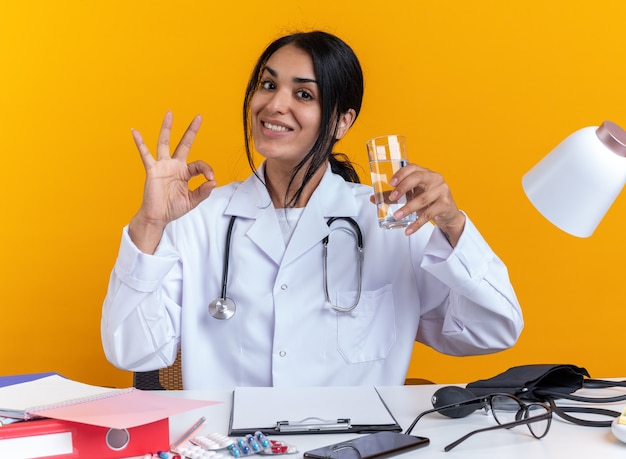 Smiling young female doctor wearing medical robe with stethoscope sits at table with medical tools holding glass of water showing okay gesture isolated on yellow background