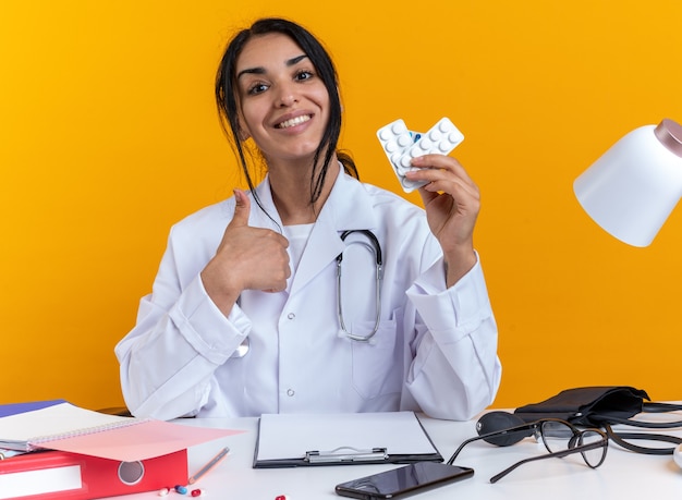 청진기가 달린 의료 가운을 입은 웃고 있는 젊은 여성 의사는 노란색 배경에 고립된 엄지손가락을 보여주는 약을 들고 있는 의료 도구를 들고 테이블에 앉아 있다