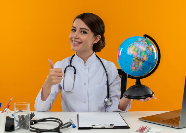 Smiling young female doctor wearing medical robe with stethoscope sitting at desk work on computer with medical tools holding globe her thumb up on isolation yellow background