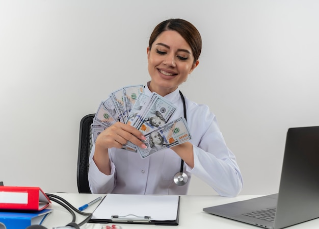 Sorridente giovane donna medico indossando abito medico con uno stetoscopio seduto alla scrivania lavora sul computer con strumenti medici tenendo e guardando in contanti con spazio di copia