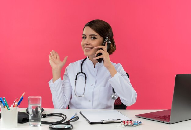 Smiling young female doctor wearing medical robe with stethoscope sitting at desk work on computer with medical tools speaks on phone and points with hand to side on pink wall
