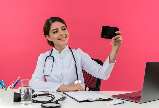 Smiling young female doctor wearing medical robe with stethoscope sitting at desk work on computer with medical tools take selfie with copy space