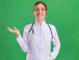 Free photo smiling young female doctor wearing medical robe with stethoscope spreading hand isolated on green wall