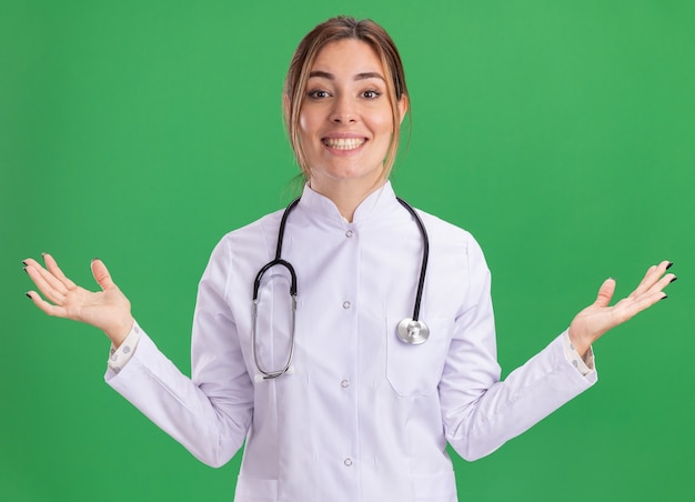 緑の壁に分離された手を広げて聴診器で医療ローブを着て笑顔の若い女性医師