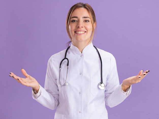 Sorridente giovane medico femminile che indossa abito medico con lo stetoscopio diffondendo le mani isolate sulla parete viola