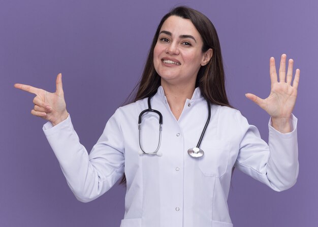 Улыбающаяся молодая женщина-врач в медицинском халате со стетоскопом стоит с поднятой рукой