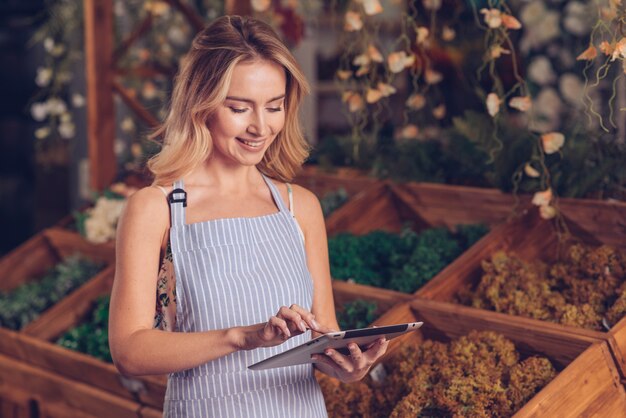 Smiling young female florist in apron touching the screen of digital tablet