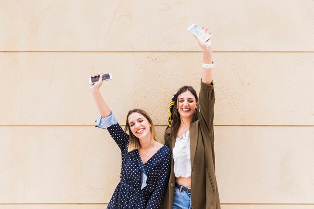 Smiling young female friends raising their hand holding mobile phones