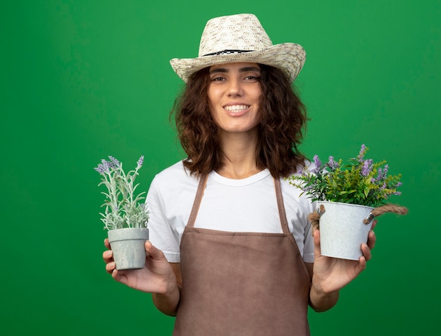 Бесплатное фото Улыбающаяся молодая женщина-садовник в униформе в садовой шляпе держит цветы в цветочных горшках, изолированных на зеленой стене