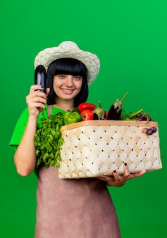 野菜のバスケットを保持しているガーデニング帽子を身に着けている制服を着た若い女性の庭師の笑顔