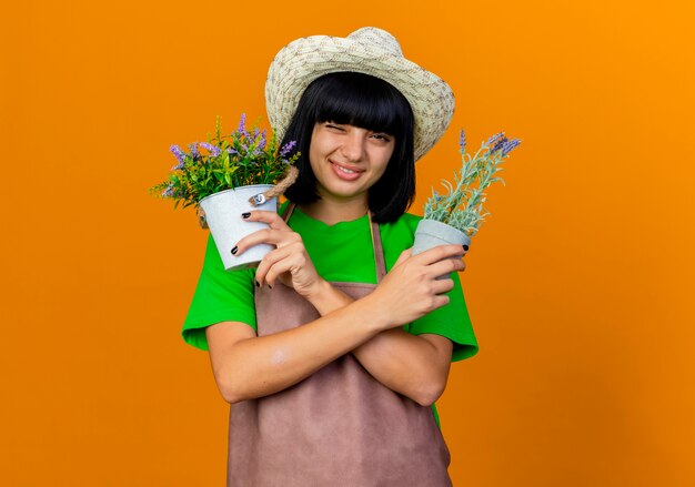 Smiling young female gardener in uniform blinks eye holding flowerpots