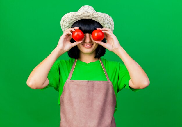 원예 모자를 쓰고 제복을 입은 웃는 젊은 여성 정원사는 토마토로 눈을 가립니다