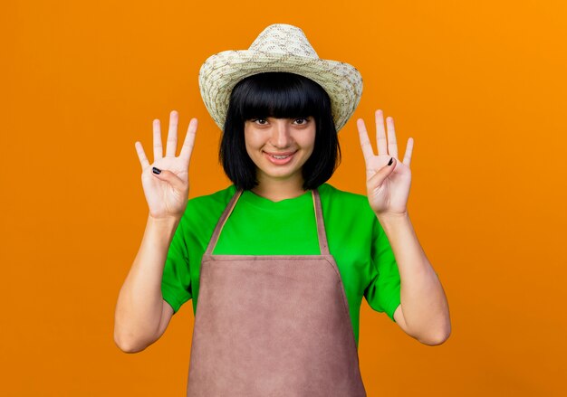 Smiling young female gardener in uniform wearing gardening hat gestures eight with fingers