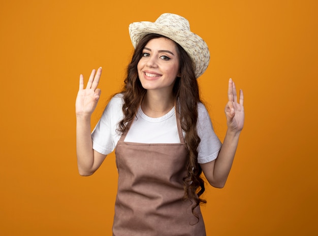 Smiling young female gardener in uniform wearing gardening hat gestures ok hand sign with two hands isolated on orange wall with copy space