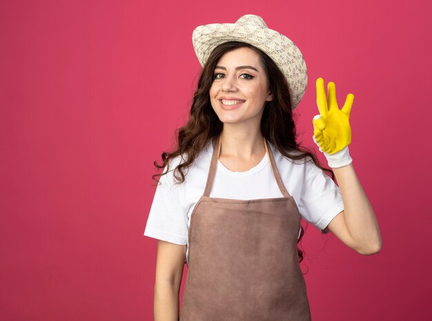 Smiling young female gardener in uniform wearing gardening hat and gloves gestures ok hand sign isolated on pink wall with copy space