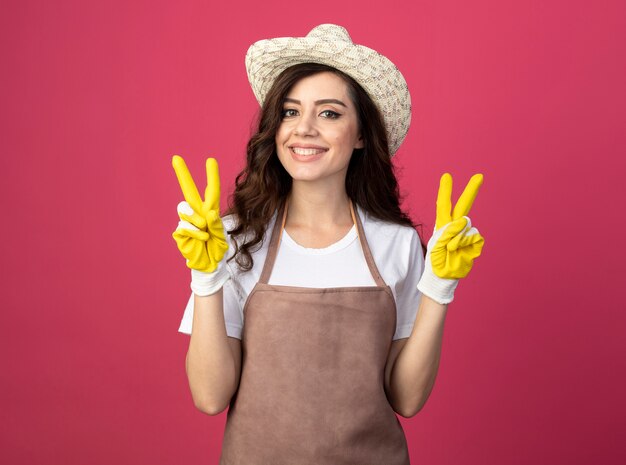Улыбающаяся молодая женщина-садовник в униформе в садовой шляпе и перчатках жесты рукой знак победы двумя руками, изолированными на розовой стене с копией пространства