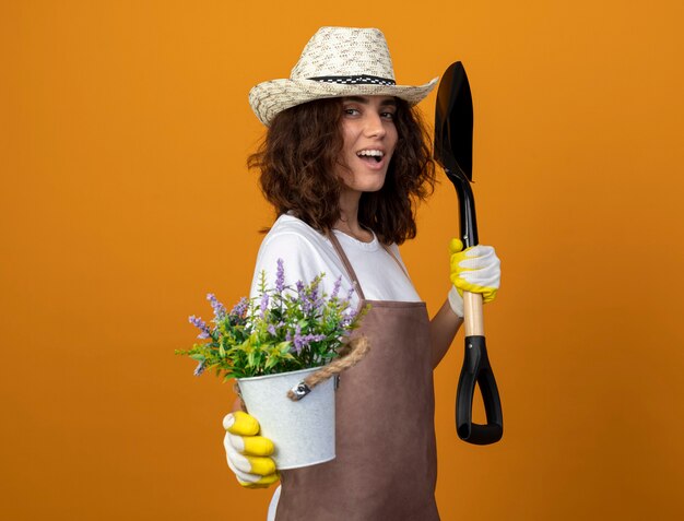 Smiling young female gardener in uniform wearing gardening hat and gloves holding out flower in flowerpot