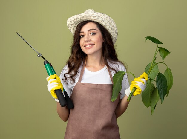 Smiling young female gardener in uniform wearing gardening hat and gloves holds plant and garden clippers isolated on olive green wall with copy space