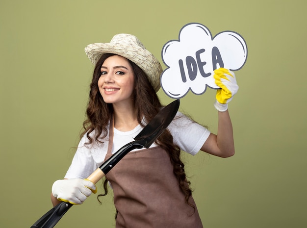 Sorridente giovane giardiniere femminile in uniforme indossando guanti e cappello da giardinaggio tiene e punta alla bolla di idea con vanga isolata sulla parete verde oliva