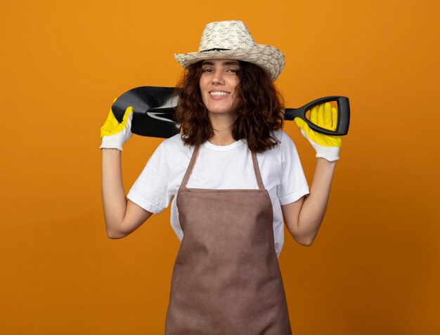 Smiling young female gardener in uniform wearing gardening hat and gloves putting spade on behind neck